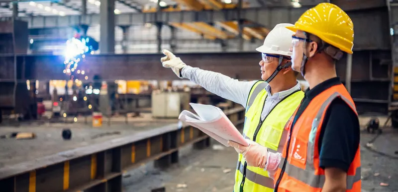 Two men in hard hats, safety glasses, and high-visibility vests standing in a steel fabrication workshop, one pointing while holding a rolled drawing, with a welder throwing sparks behind them