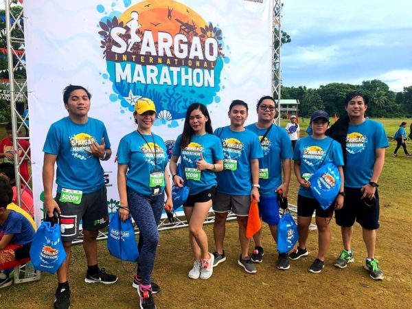 Eight runners in matching blue Siargao International Marathon shirts with race bibs and blue drawstring bags, standing on grass in front of the event backdrop under an overcast sky