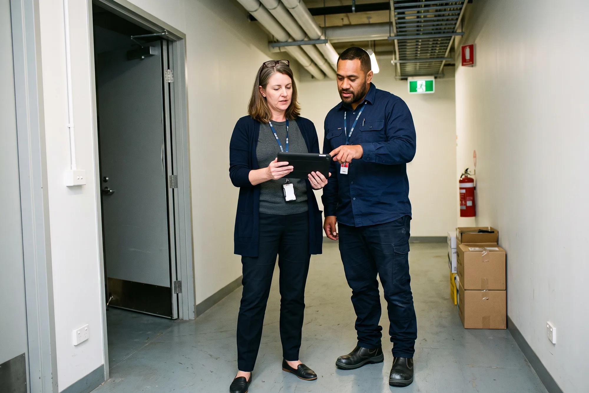 A facilities manager holding a tablet and a building services supervisor pointing at it as they talk in a plant room service corridor