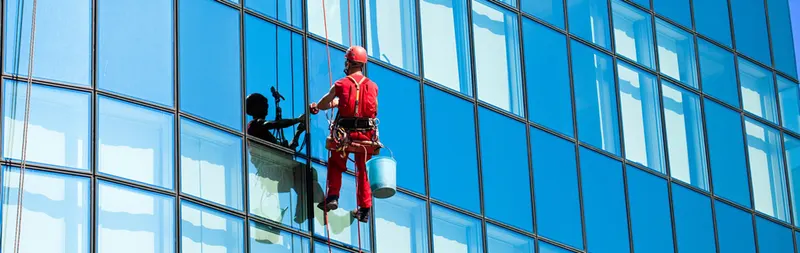 A rope access worker in red overalls and a red helmet abseiling down the glass facade of an office tower, with a blue bucket clipped to his harness