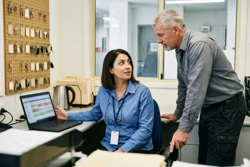A facilities coordinator at her desk in a commercial building management office turns a laptop towards an air-conditioning technician who leans in beside her, with a pegboard of keys on the wall behind them