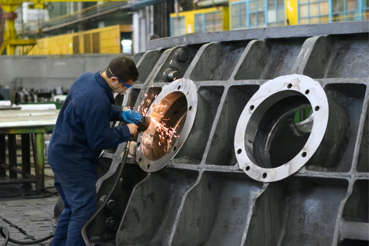 A fitter in blue overalls, safety glasses and a dust mask grinding a flange on a large cast housing in a workshop, sparks coming off the disc