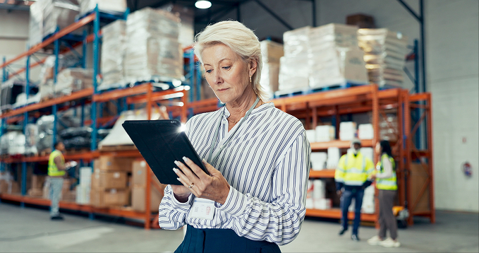A woman with short white hair in a striped shirt reads a tablet in a warehouse aisle, with stacked pallet racking behind her and two workers in high-visibility vests further down the aisle