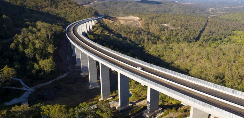 An aerial view of a curving concrete viaduct carrying a dual carriageway on tall piers across a forested valley, with a single car travelling on the near carriageway