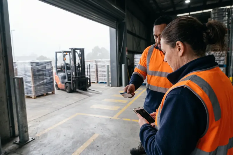 A warehouse shift supervisor in a navy fleece and orange hi-vis vest checks a phone beside a forklift driver at a loading dock door while he holds out a licence card