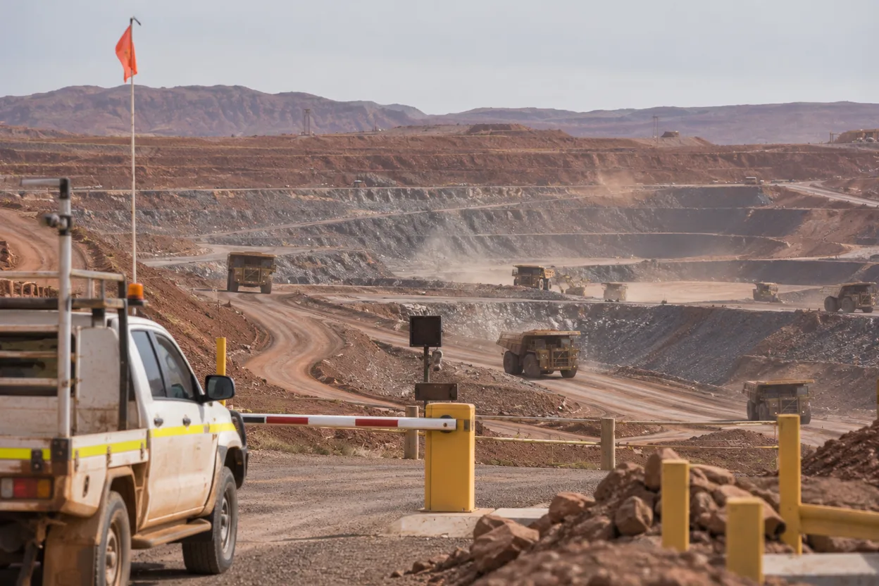 An open-cut mine pit with haul trucks working the benches, seen from the site access road near the boom gate