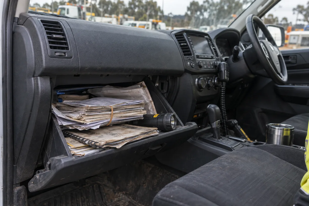 The open glovebox of a ute with a bundle of folded plant certificates and service dockets held by a rubber band, a pre-start booklet, and a torch
