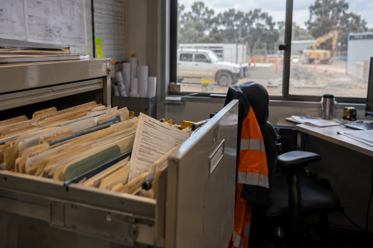 An open filing cabinet drawer of contractor personnel files in a site office, manila folders with handwritten tabs, a medical form half pulled out, everything too soft to read
